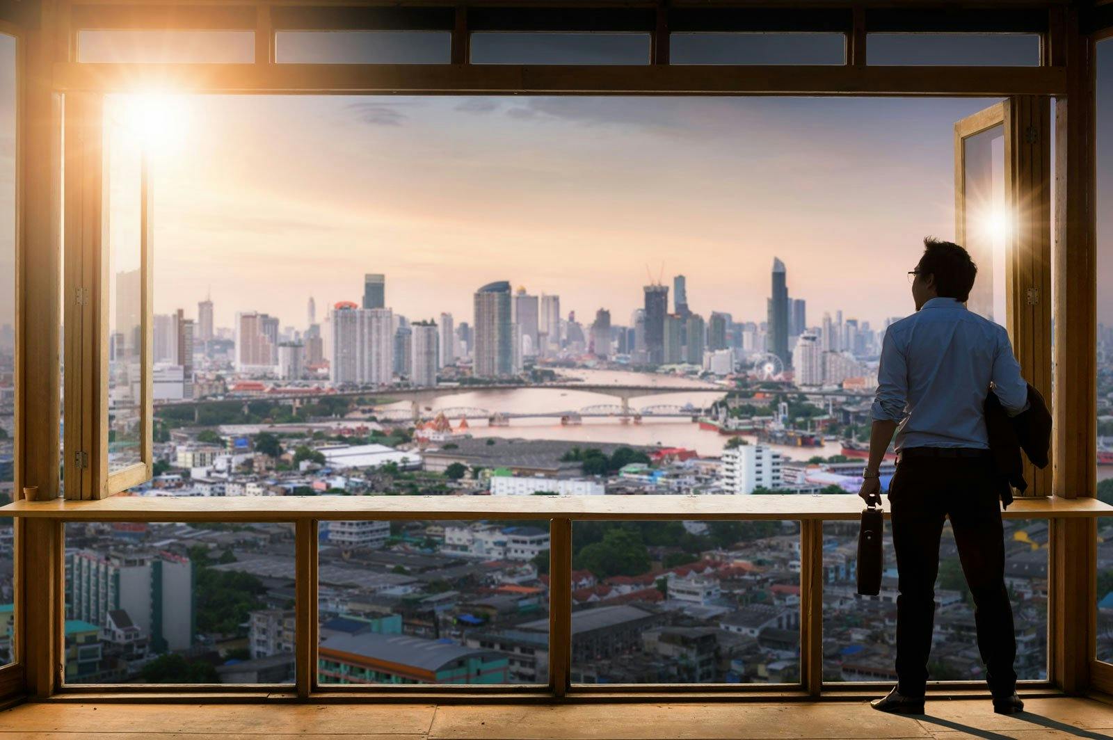 large open window with a single man standing on the right side and looking out at a view of the city